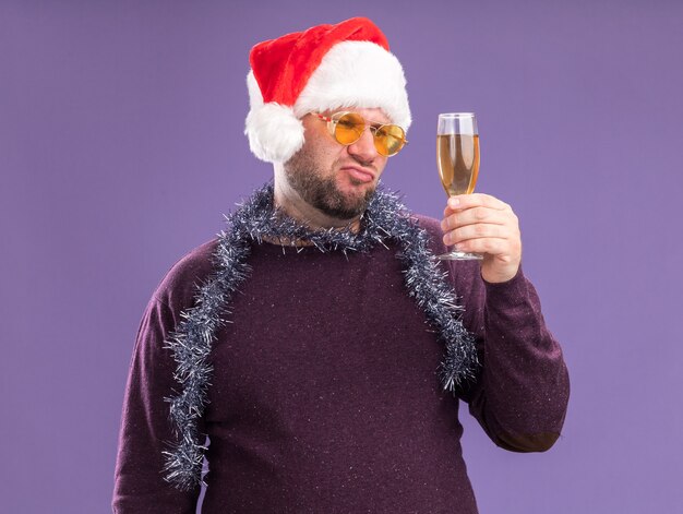 Zweifelhafter Mann mittleren Alters, der Weihnachtsmütze und Lametta-Girlande um den Hals mit Gläsern trägt, die Glas des Champagners halten und betrachten, lokalisiert auf lila Wand