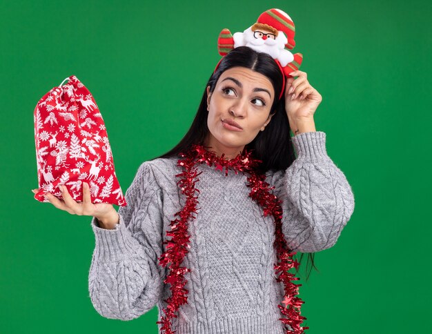 Zweifelhaftes junges kaukasisches Mädchen, das Weihnachtsmann-Stirnband und Lametta-Girlande um den Hals hält, der Weihnachtsgeschenksack berührt, der Stirnband berührt, das Seite lokalisiert auf grüner Wand betrachtet