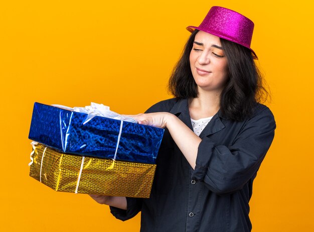 Zweifelhaftes junges kaukasisches Partymädchen mit Partyhut, das Geschenkpakete isoliert auf oranger Wand hält und betrachtet
