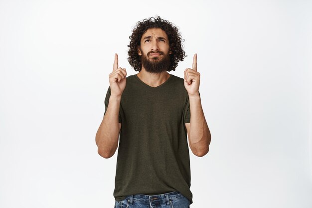 Zweifelnder Mann, der mit dem Finger nach oben zeigt und mit zögerndem skeptischem Gesichtsausdruck vor weißem Hintergrund steht