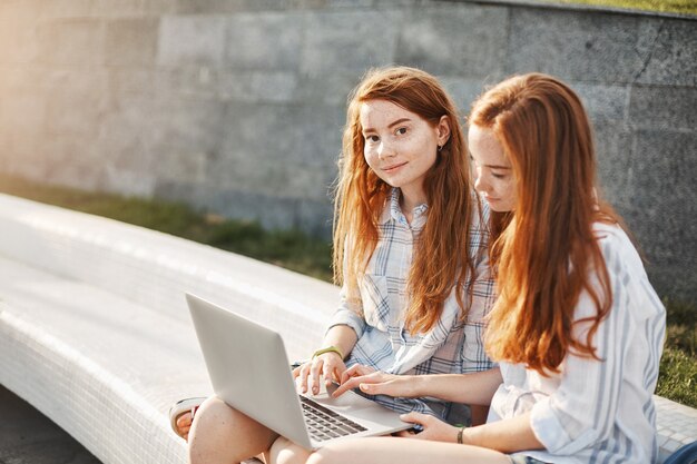 Zwillinge starten Geschäftsprojekt. Porträt von entspannten und niedlichen weiblichen Stundents mit Ingwerhaar und Sommersprossen, sitzend im Stadtplatz, Arbeiten mit Laptop