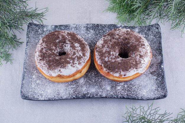 Kostenloses Foto zypressenblätter um eine mit mehl bedeckte platte mit zwei donuts oben auf weißem hintergrund.