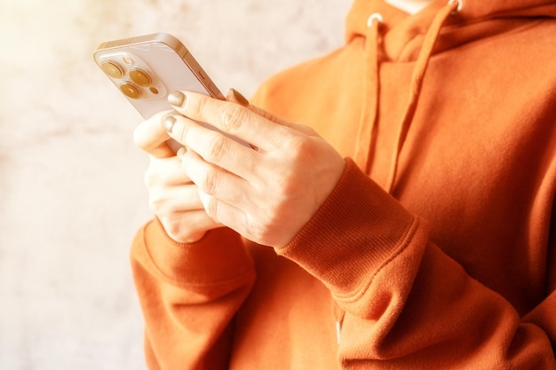 03042022 Manos femeninas de Moscú sosteniendo un teléfono inteligente y enviando un mensaje de texto a una mujer joven sin rostro en una sudadera con capucha marrón naranja y usando un teléfono móvil en un fondo gris cerca de una bengala