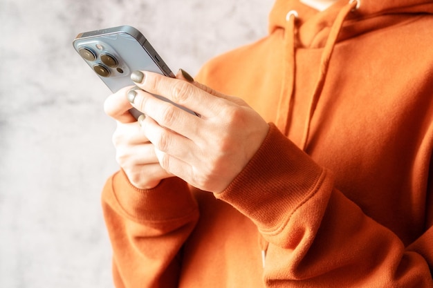 03042022 Moscú manos femeninas sosteniendo un teléfono inteligente y enviando un mensaje de texto a una joven sin rostro con capucha marrón naranja y usando un teléfono móvil en un fondo gris de cerca