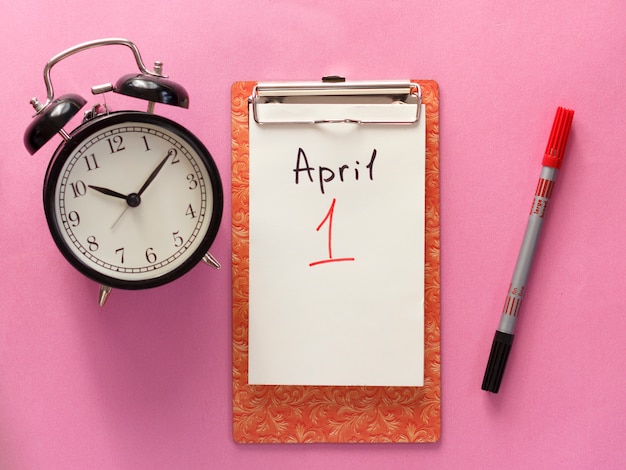 Foto 1 de abril día del tonto, cuaderno, reloj, pluma. aplanado sobre fondo rosa.
