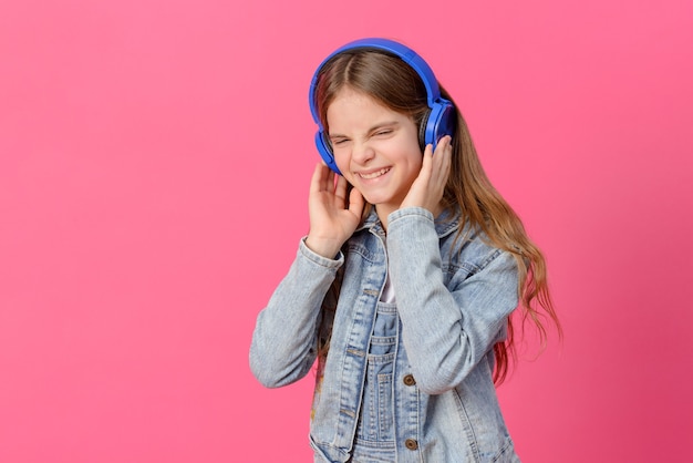 1 linda garota branca de 10 anos em uma jaqueta jeans azul com fones de ouvido em uma superfície rosa