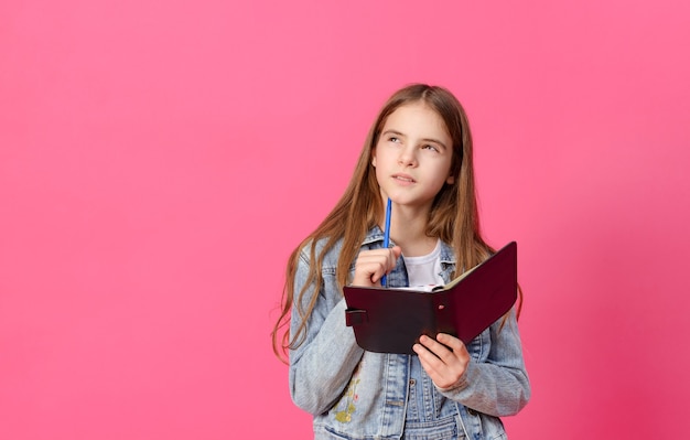 1 linda garota branca pensativa de 10 anos de idade em uma jaqueta jeans azul com um caderno e uma caneta em uma superfície rosa