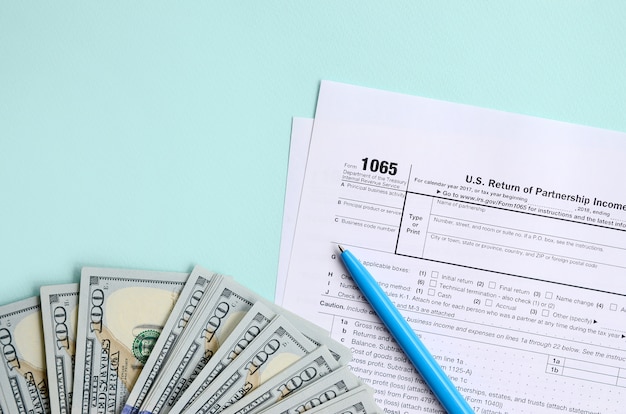 1065 Steuerformular liegt nahe hundert Dollarscheinen und blauem Stift auf einem hellblauen Hintergrund. US-Rückkehr für Elterneinkommen