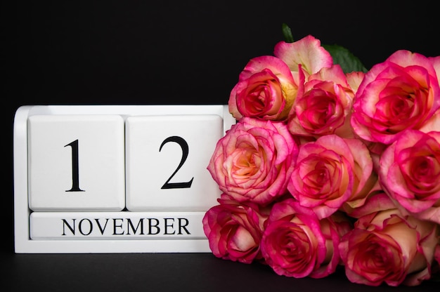 12. November Holzkalender, weiß auf schwarzem Hintergrund, rosa Rosen liegen in der Nähe. Postkarte, Raum.