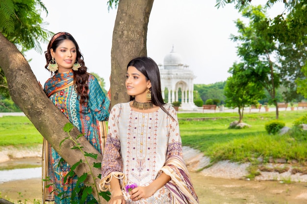 2 chicas atractivas con vestidos tradicionales y joyas para una sesión de fotos de moda en un jardín con un árbol