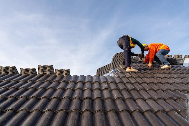 2 Dachdecker in Schutzkleidung und Handschuhen mit Luft- oder pneumatischer Nagelpistole und Installation von Betondachziegeln auf dem neuen Dachkonzept des im Bau befindlichen Wohngebäudes