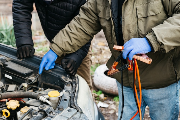 2 Ingenieros mecánicos que cargan la batería del automóvil con electricidad utilizando cables de puente en exteriores. Cables de puente rojos y negros en manos masculinas del mecánico de automóviles. Mans en guantes trabajando en la estación de servicio de reparación de automóviles