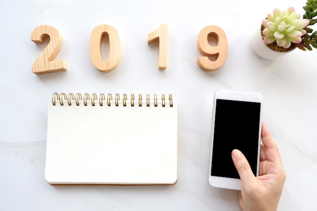 2019 letras de madeira, mão segurando o telefone inteligente com tela em branco e papel de caderno em branco