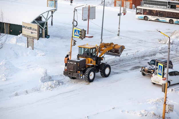 23.01.2023, Kemerovo, Rusia. El tractor naranja limpia la nieve de la carretera y la carga en un camión