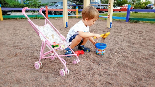 3 Jahre alter Kleinkindjunge, der mit Kinderwagen für Puppen auf dem Spielplatz spielt