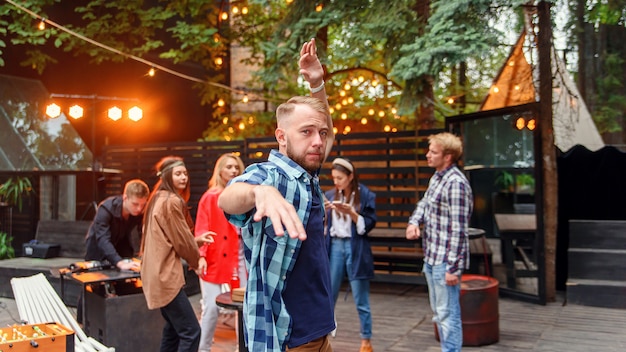 30s caucasiano jovem bonito dançando perto da câmera no fundo de seus amigos na festa no acolhedor jardim da noite.