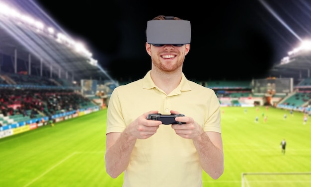 3D-Technologie, Virtual Reality, Sport, Unterhaltung und Personenkonzept - Mann mit Virtual-Reality-Headset oder 3D-Brille, der mit Gamecontroller-Gamepad über Fußballfeld auf Stadionhintergrund spielt