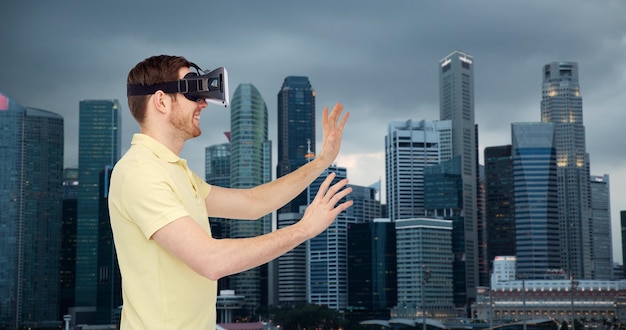 3D-Technologie, virtuelle Realität, Reise-, Unterhaltungs- und Personenkonzept - glücklicher junger Mann mit Virtual-Reality-Headset oder 3D-Brille, der ein Spiel über dem Hintergrund der Wolkenkratzer der Stadt Singapur spielt