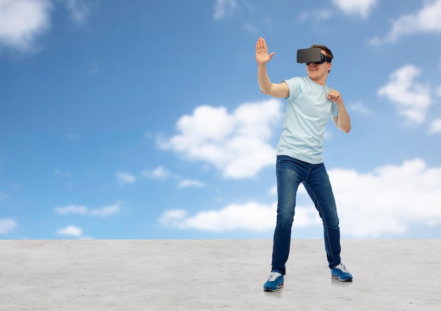 3D-Technologie, virtuelle Realität, Unterhaltung, Cyberspace und People-Konzept - junger Mann mit Virtual-Reality-Headset oder 3D-Brille, der ein Spiel spielt und über blauem Himmel und Wolkenhintergrund kämpft
