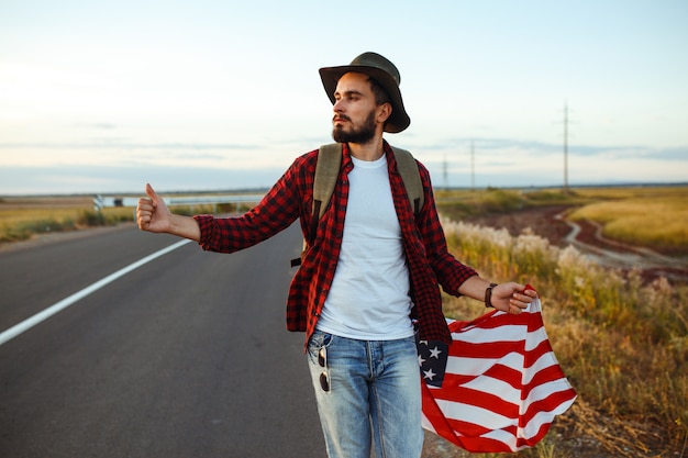 4 de julho. Quatro de julho. Americano com a bandeira nacional. Bandeira americana. Dia da Independência. Feriado patriótico. O homem está usando chapéu, mochila, camisa e calça jeans.