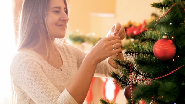 4 imágenes de 4 k de hermosa mujer sonriente con el pelo largo colgando adornos y guirnaldas en el árbol de Navidad en la sala de estar. Familia preparando y decorando la casa en vacaciones y celebraciones de invierno.