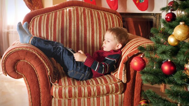 4 imágenes de k de niño pequeño en suéter de lana acostado en un gran sillón junto al árbol de Navidad y usando tableta digital. Niño divirtiéndose en celebraciones y vacaciones de invierno.