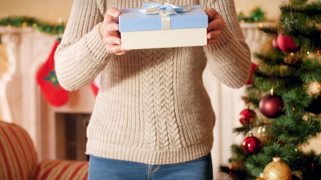 4 k imagens de jovem com uma camisola de lã em pé ao lado da árvore de Natal e lareira e mostrando a caixa de presente de Natal com fita e arco.