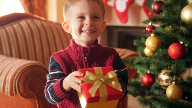 4 k imagens de menino feliz e sorridente segurando o presente de natal na caixa e mostrando seu presente na câmera. criança recebendo presentes e presentes do papai noel nas comemorações e feriados de inverno