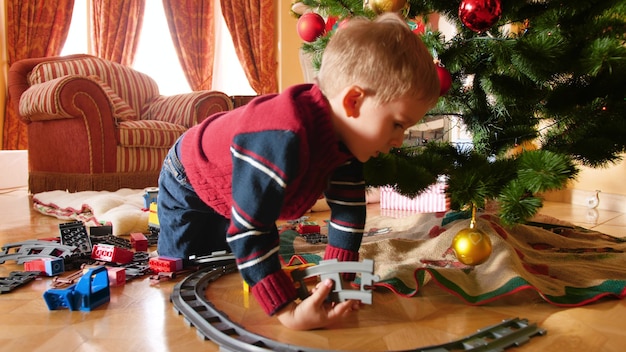4K-Aufnahmen des kleinen Jungen erhielten Spielzeugeisenbahn und Zug zu Weihnachten vom Weihnachtsmann. Kind baut Schienen um Weihnachtsbaum im Wohnzimmer
