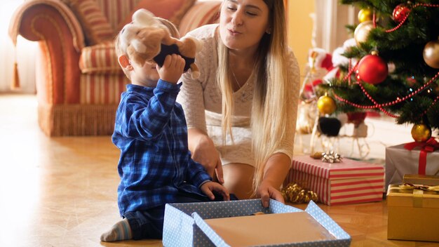 4K-Aufnahmen von einem entzückenden kleinen Jungen, der seine Geschenkbox vom Weihnachtsmann öffnet und einen Spielzeugplüschhund herausnimmt. Familie, die an Winterferien und -feiern Geschenke gibt und empfängt.