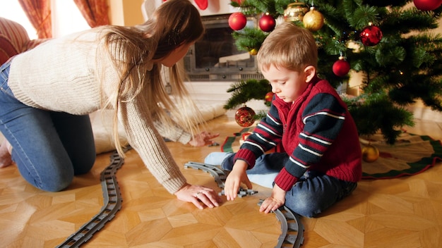 4K-Aufnahmen von glücklich lächelnder Mutter, die ihrem Sohn beim Bau einer Eisenbahnstrecke aus Ziegeln unter dem Christams-Baum hilft. Kind erhält Geschenke und Geschenke vom Weihnachtsmann an Winterferien und an Feiertagen