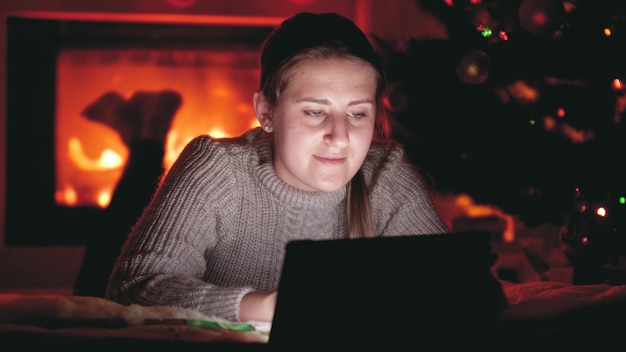 4K-Aufnahmen von lächelnden Mädchen, die auf einem Tablet im Internet surfen, während sie neben einem Kamin und einem leuchtenden Weihnachtsbaum liegen