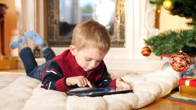 4k Nahaufnahmen von kleinen Jungen mit digitaler Tablette, die auf dem Boden im Wohnzimmer neben Weihnachtsbaum und Kamin liegt. Kinder, die Spaß an Winterferien und Feiern haben.