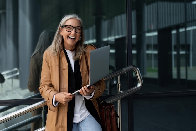 50-jährige erfolgreiche Geschäftsfrau mit einem Laptop in ihren Händen lächelt breit außerhalb des Büros