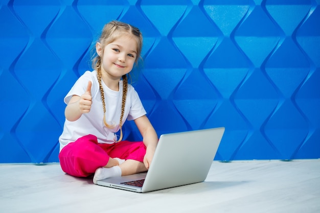 7 Jahre altes Mädchen in einem weißen T-Shirt sitzt mit einem Laptop auf dem Boden und drückt die Tasten