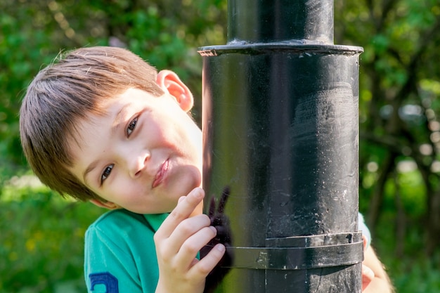 8-jähriger Junge mit dunklen Haaren steht hinter einem Laternenpfahl aus Metall. Netter Brunetjunge, der hinter einem Laternenpfahl Verstecken spielt. Kinder spielen im Park