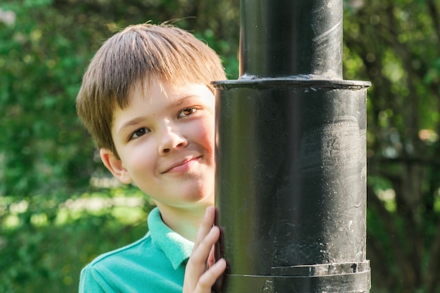 8-jähriger Junge mit dunklen Haaren steht hinter einem Laternenpfahl aus Metall. Netter Brunetjunge, der hinter einem Laternenpfahl Verstecken spielt. Kinder spielen im Park