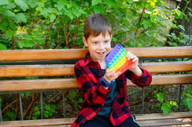 8-jähriger Junge spielt mit Popit im Park. glückliches Kind mit Spielzeug. Kind trägt helle Sommer Freizeitkleidung. mehrfarbige Pop-It-Nahaufnahme. Anti-Stress-Spielzeug aus Kunststoff.