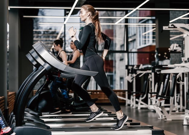 A atlética garota vestida com uma roupa esporte preta, correndo na esteira no ginásio moderno.
