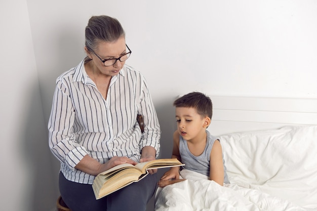 A avó de óculos e camisa branca lê um livro para o neto deitado na cama