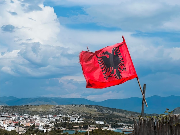 A bandeira nacional da Albânia ondulando contra o belo céu e a aldeia montanhosa