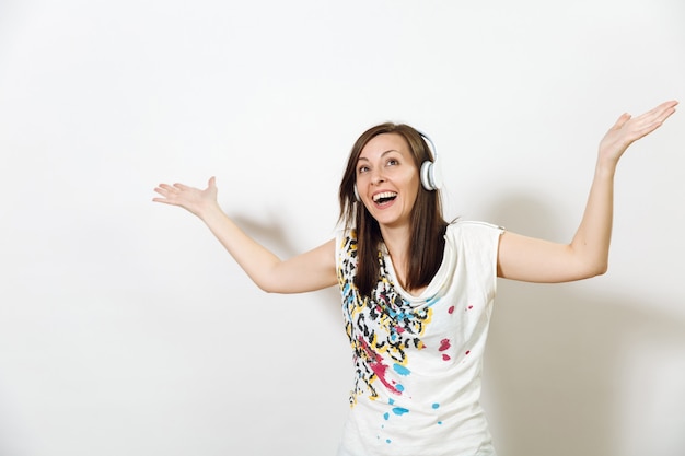 A bela e emocional jovem europeia de cabelos castanhos feliz com pele saudável e limpa e sorriso encantador, vestida com roupas leves e casuais, ouve e curte música em fones de ouvido em um background branco