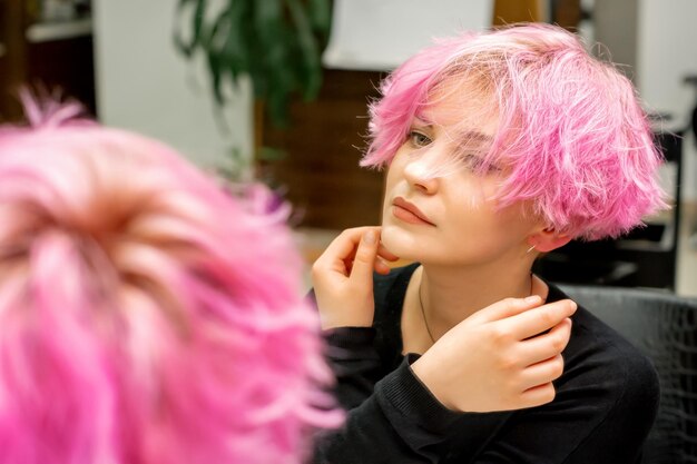 A bela jovem mulher caucasiana com um novo penteado rosa curto olhando seu reflexo no espelho verificando o penteado em um salão de cabeleireiro