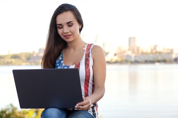 A bela jovem senta-se no parque perto do rio com o laptop.