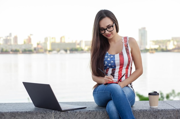 A bela jovem senta-se no parque perto do rio com o laptop.