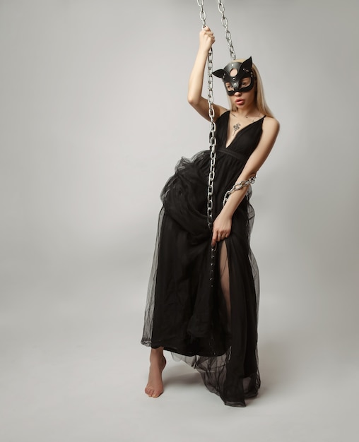 A bela loira com uma máscara de gato em um vestido de noite sexy em um fundo branco posando com correntes de metal