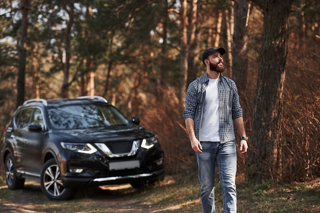 A bela luz do sol entra pelas árvores. Homem barbudo perto de seu carro preto novo na floresta. Conceito de férias