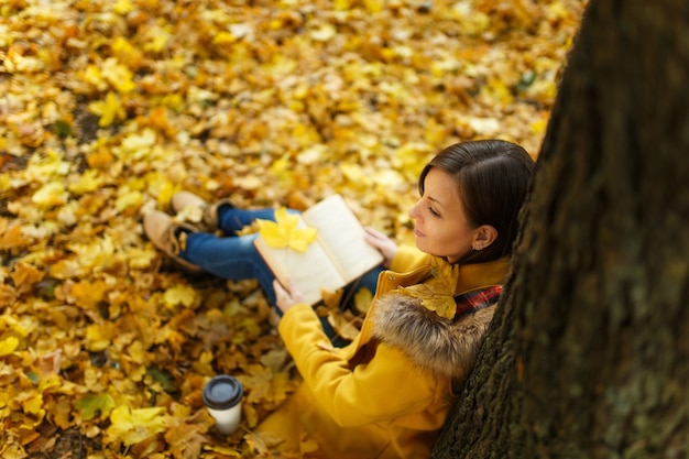 A bela mulher de cabelos castanhos feliz com casaco amarelo e calça jeans, sentado sob a árvore de bordo com uma xícara de chá ou café e um livro vermelho no parque da cidade de outono. Folhas de outono dourado. Conceito de leitura. Vista do topo.