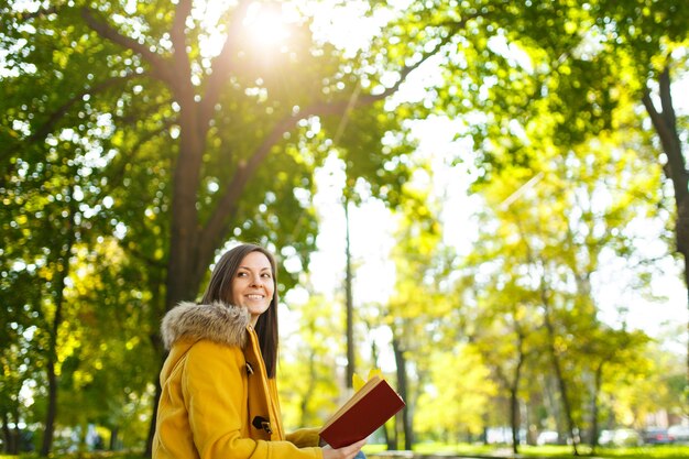 A bela mulher de cabelos castanhos feliz com um casaco amarelo senta-se sozinha no parque e lê um livro no dia quente de outono. Folhas de outono amarelas.