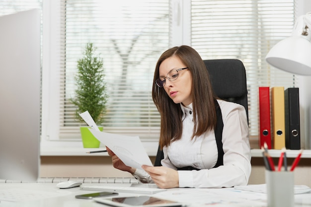 A bela mulher de negócios de cabelo castanho, séria e absorta, de terno e óculos, sentada à mesa com um tablet, trabalhando no computador com um monitor moderno com documentos em um escritório leve, olhando para o lado